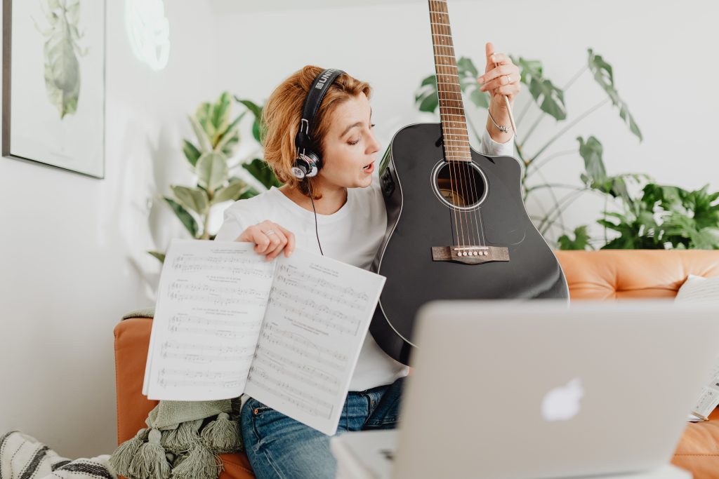 A woman with headphones teaches guitar online, holding sheet music and an acoustic guitar.
